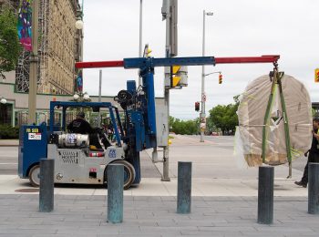 Photo couleur montrant un petit chariot élévateur transportant une grande pierre ovale et plate suspendue à un bras, en milieu urbain.