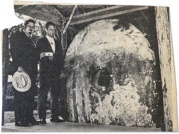 Coupure de presse d’une photo en noir et blanc. Deux hommes en costume sont debout sur une plateforme, sous un petit toit, à côté d’un grand disque de pierre ovale avec un trou au milieu.