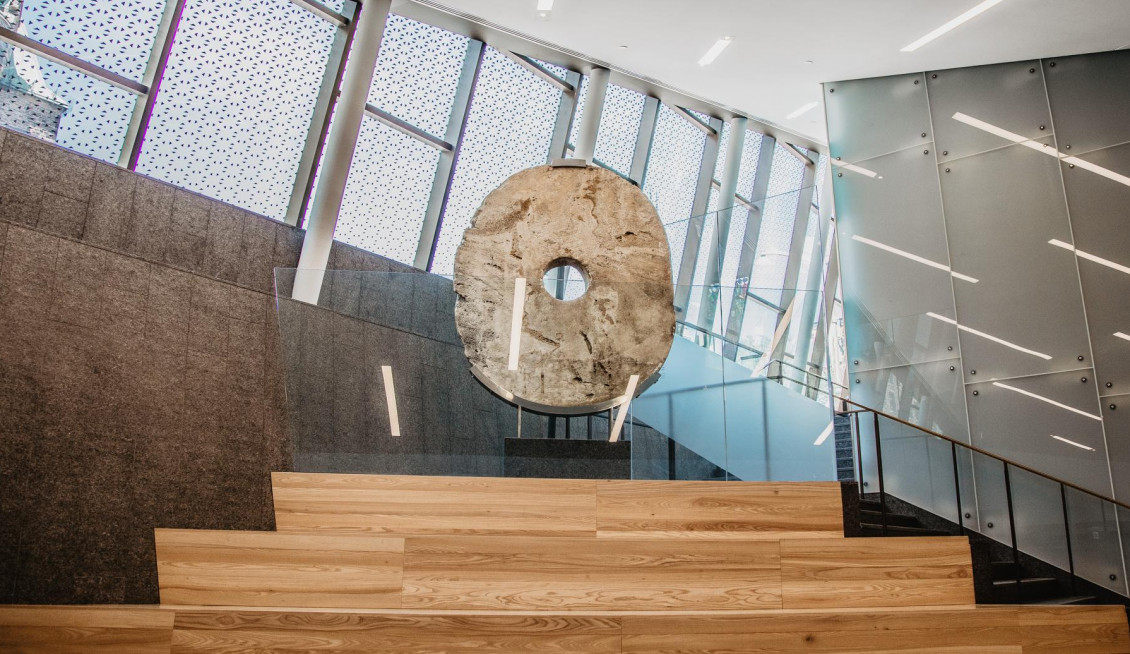 Grande pierre plate, taillée en forme ovale avec un trou au centre, et posée sur un socle en haut d’un large escalier en bois, dans un atrium.